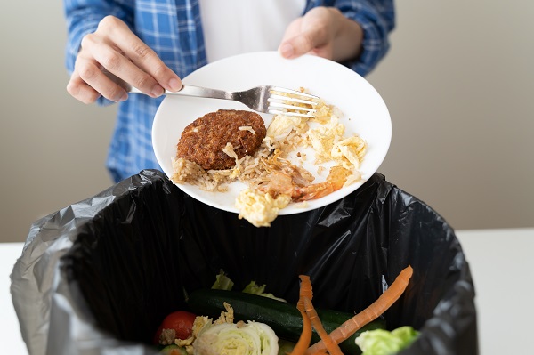 A person scraping food from a plate into a trash can with other food scraps.
